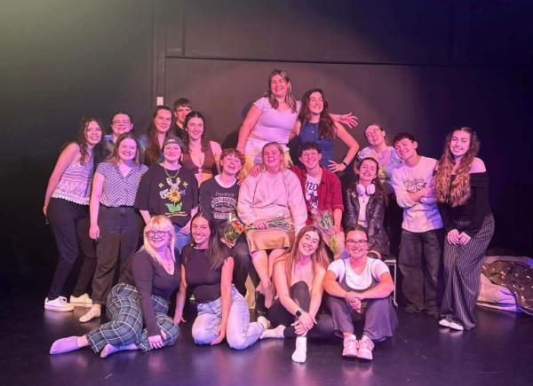 A group of musical theatre society members clump together for group photo, under a spotlight, against a black wall