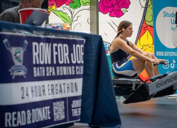student in blue and navy rowing kit rows on a static rowing machine in the background. The foreground shows a banner reading 'Row for Joe' 